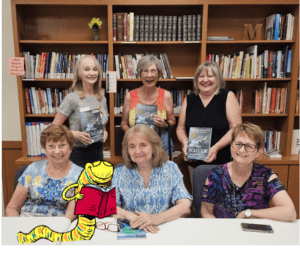 A group of six women smiling in a library, holding books.
