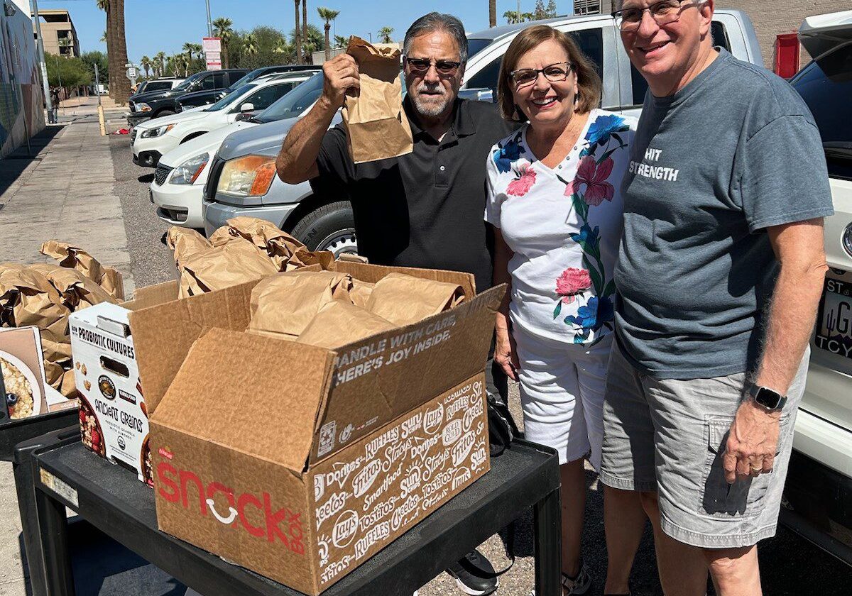 Three people smiling by a table with boxes outdoors on a sunny day.