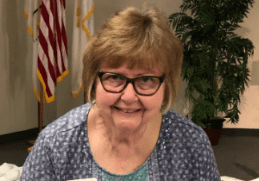 Smiling elderly woman wearing glasses and a name tag indoors.